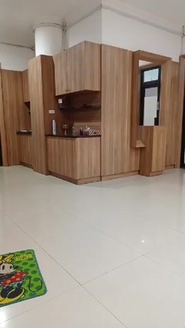 The video shows an interior space, likely a kitchen or breakroom, with light-colored wood cabinets arranged against the walls. The floor is tiled in a glossy, off-white color. A small, green rug with cartoon characters is visible in the lower left corner. The lighting is bright, suggesting the presence of artificial lights. The room appears clean and well-maintained. No people are visible in the video.