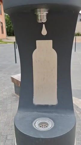 This is a close-up video of a public water fountain, likely in a park or public square, given the paved ground and a bench visible in the background. The fountain itself is a dark gray, modern-looking structure with a stylized outline of a bottle and a water drop printed on its front. A metal spout at the top dispenses water, which flows down into a drain at the base. The action captured is the water flowing from the spout, suggesting it is being used, or perhaps it's a demonstration of its function. The lighting suggests it's daytime, and the overall scene appears to be outdoors. The video is shot from a low angle, focusing on the fountain itself and the water flow. There are no people visible in the frame.