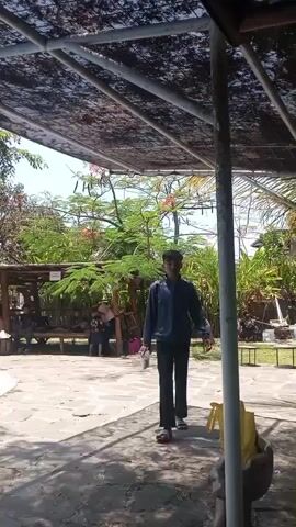 A video shows a young man standing near a swimming pool under a shaded area. In the background, people are seated under a thatched-roof shelter. Trees and foliage surround the area, with some red flowers visible on a tree. The ground is paved with stone tiles. The overall atmosphere appears to be a leisure or recreational setting.
