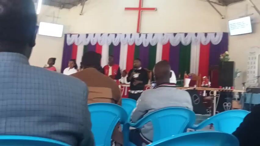The video appears to show the interior of a church or meeting hall. Several people are visible, some seated on blue plastic chairs. A colorful backdrop with vertical stripes of red, white, green, and purple is positioned behind the group. There's also a white decorative element along the bottom edge of the backdrop. A speaker and other equipment can be seen in the background. The video seems to be taken from a slightly elevated position, perhaps from someone sitting down, and the perspective is upside down.