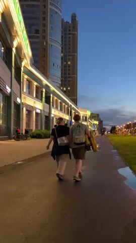 The video shows a low-angle, medium shot from behind, following two individuals, a man and a woman, as they walk hand-in-hand along a wet, paved sidewalk. The woman is wearing a dark shirt, light-colored pants, and carrying a light-colored crossbody bag. The man is wearing a light-colored t-shirt, dark shorts, and a light-colored backpack. He also has a yellow jacket or bag draped over his right shoulder. They are walking away from the camera, towards a tall, modern building with many windows. To their left is a long, low-rise building with illuminated storefronts. The sky is a deep blue, indicating dusk or early evening. To the right of the sidewalk is a patch of green grass with some puddles of water. The scene appears to be an urban environment.