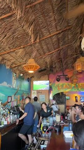The video shows a television screen displaying a soccer match with the score ESP 1 ARG 0 at 129 minutes and 59 seconds. The television is mounted on a wall within a bar or restaurant that features a thatched roof and tropical-themed decor, including a colorful mural with a parrot and palm trees. The camera pans to show a crowded bar area with people watching the game. Several individuals are visible at the bar and standing around, engaged in watching the match. The atmosphere appears lively, suggesting a sporting event is being watched by a group of patrons.