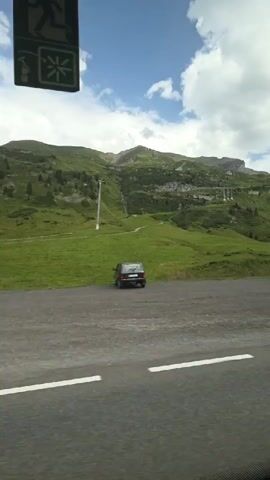 The video shows a view from a vehicle traveling along a paved road with white lane markings. The road is in the foreground. Beyond the road is a grassy slope leading up to a mountain. The mountain is covered in green grass and trees, and there are some structures, possibly ski lifts, visible on its side. The sky is blue with white, puffy clouds. A few cars are parked or driving on the road and in the background. The overall scene is a scenic mountain landscape on a partly cloudy day.