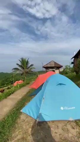 The video shows a campsite on a grassy hill overlooking the ocean. There are several tents, some red and some blue, pitched along a concrete path. A small gazebo stands near the tents. A palm tree is visible near the blue tent. The ocean is calm and blue, and the sky is partly cloudy.