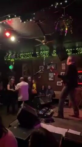 The video shows a live band performing in a dimly lit, crowded venue. The band is on a small stage, with a guitarist visible in the foreground, playing with energy and enthusiasm. In front of the stage, several people are dancing, including two couples. One woman is wearing a yellow shirt, while one man is wearing a white polo shirt. The walls are brick and adorned with framed pictures and string lights, creating a festive atmosphere. The camera work is shaky, offering a slightly off-kilter perspective of the scene.