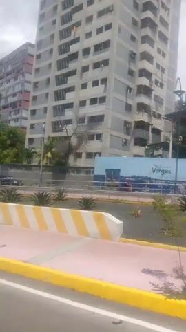 The video shows a view from a moving vehicle, looking towards a street lined with trees and tall buildings. In the foreground, there is a sidewalk with a red and white patterned surface, bordered by a yellow stripe. Beyond the sidewalk is a strip of green grass, with a few scattered bushes and a small brown rock. Several trees with green foliage are visible. Behind the trees, a white metal fence runs along the edge of the property. Across the street, there are several cars, including a white sedan and a darker colored vehicle. The background is dominated by a very tall, multi-story building with many windows. The building appears to be light colored with some pink or red accents. The sky is overcast with some lighter patches. The video is shaky, indicating it was filmed from a moving vehicle.