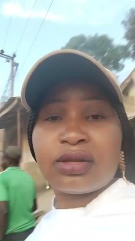 A woman wearing a white shirt and a beige baseball cap is taking a selfie video outdoors. She has braids and is wearing gold earrings.  There are two men in the background, one wearing a pink shirt and the other a green shirt. The environment appears to be a residential area with buildings and trees visible. There are utility poles overhead.