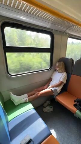 A blonde woman with sunglasses on is sitting on a train facing a window. She is wearing a white t-shirt, denim shorts, and white socks with red accents. Her legs are crossed and extended, resting on a blue and green seat. Her shoes are placed on the floor in front of her. The train appears to be moving, as the trees outside the window are blurred. The interior of the train car has a light-colored ceiling with yellow accents, and multi-colored seats.