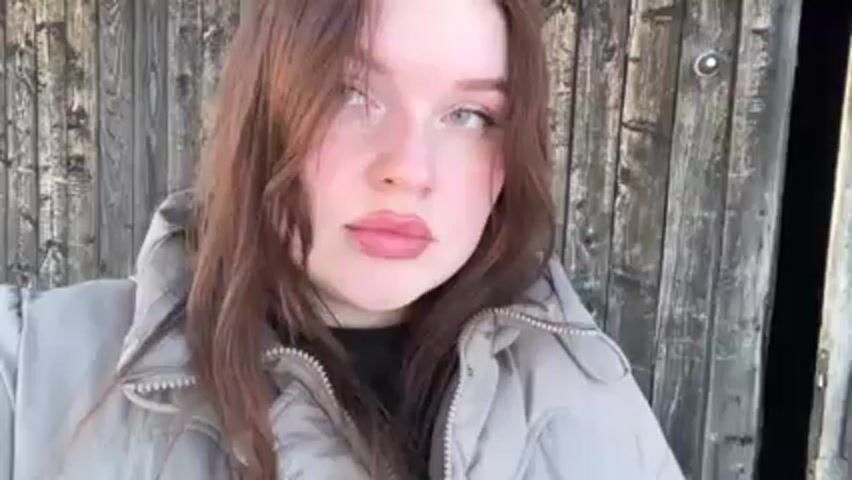 A young woman with long brown hair and light skin is standing outside in front of a weathered wooden structure, possibly a shed or barn. She's wearing a light gray padded jacket. She briefly closes her eyes and then opens them, looking to the side and then upward. She runs her hand through her hair. The light is bright and seems to be coming from above.
