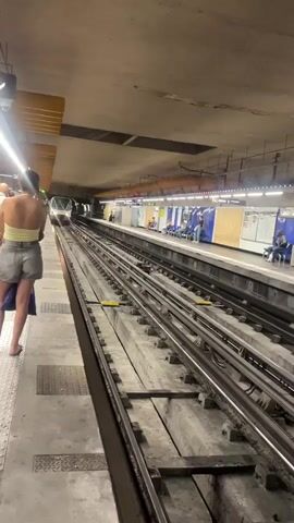 A train is approaching a subway station platform. A person is standing on the platform facing the train, with their back to the camera. The person is wearing a light-colored tank top and denim shorts. Other people are visible sitting on benches on the platform across the tracks. The station appears to be underground, with a concrete ceiling and lighting fixtures. The train tracks are in the foreground.