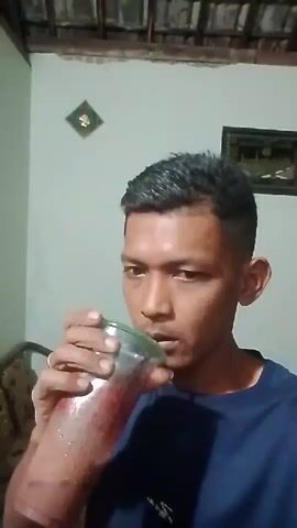 A young man with short dark hair and a dark blue t-shirt is drinking a reddish-brown beverage from a clear plastic cup with a green lid. He is holding the cup with his right hand and appears to be at home, with a tiled ceiling and a wall with some framed artwork visible in the background. The man is looking towards the camera.