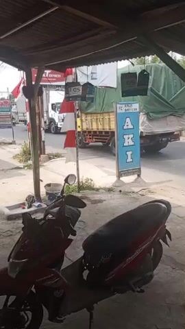 The video shows a street scene, likely in Southeast Asia, with a focus on a roadside shop. In the foreground, two motorcycles are parked under a corrugated metal roof structure. One is a red Honda. Beyond the motorcycles, a busy street is visible. A truck with a green tarp covering its cargo is passing by, followed by a silver pickup truck. Further down the street, there is a sign that reads "AKI" and "GS BATTERY", indicating a battery shop. Several GS battery advertisements are hanging. A person on a motorcycle wearing a helmet is riding past the battery shop. In the distance, other vehicles and buildings are visible. The environment appears to be a mixed commercial and residential area with a mix of modern and older infrastructure. The overall impression is one of a typical day in a bustling urban or semi-urban environment.