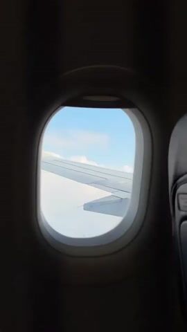 The video shows a view through an airplane window. The wing of the plane is visible, with clouds and a blue sky beyond. The interior of the plane is dark around the window.