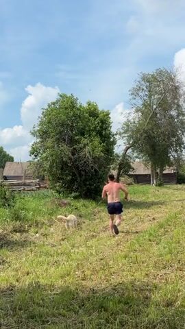 A man and a dog run up a grassy slope toward a cluster of rustic wooden buildings under a blue sky with scattered clouds. The man, wearing dark blue shorts, is shirtless and his back is to the camera. The dog, a light-colored canine, runs slightly ahead of him. Dense green trees flank the slope. The ground is covered in short, cut grass.