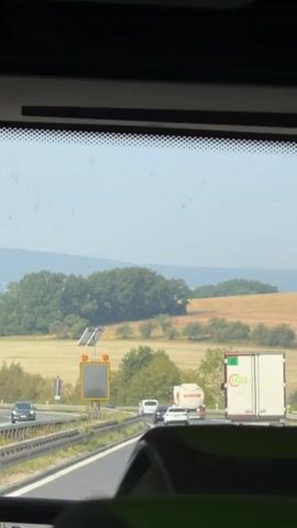 The video shows a view from inside a vehicle looking out through the windshield at a highway. There are multiple vehicles on the highway, including cars, a tanker truck, and a large white truck with a green logo. The road curves to the left. Beyond the highway, there are fields and trees, with rolling hills in the distance under a clear blue sky. The sun appears to be shining, casting light on the landscape. There is a yellow sign with a reflective panel on the side of the highway. The interior of the vehicle, including part of a seat, is visible in the foreground.