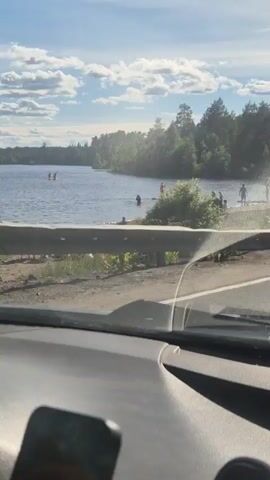 The video captures a lakeside scene from inside a vehicle. The vehicle is moving, and the camera pans across the view. On the left side of the frame, a body of water is visible with several people in the distance, some appearing to be on paddleboards. Closer to the shore, more people are gathered, some standing in the water and others on the grassy bank. A white SUV is parked near the water's edge. On the right side of the frame, a dark-colored SUV is also visible, parked or driving on a paved area adjacent to the road. Further down the road, several other cars are parked in a lot. The scene is set on a sunny day with a blue sky and scattered clouds, surrounded by trees. The interior of the vehicle, including the dashboard and windshield, is partially visible in the foreground.