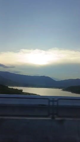 A time-lapse video shows a body of water surrounded by mountains under a partly cloudy sky. The sun is visible through the clouds, casting a bright light. The camera is positioned on a bridge, with a railing visible in the foreground. The video appears to be shot during the late afternoon or early evening, as indicated by the lighting conditions.