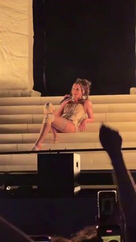 A woman with braided hair sits on a wide, cream-colored staircase. She is wearing a light-colored, strapless corset-style top and light-colored shorts. She has white knee-high socks or stockings on both legs, and she is wearing light-colored shoes. Her left leg is bent at the knee, and her left arm is resting on her bent leg. Her right leg is extended in front of her, and her right arm is resting on the step beside her. She is looking up and to her left with a smile. Behind her is a large, dark screen displaying a vertical, abstract image with red and white streaks. In the foreground, there are hands and cell phones visible, suggesting an audience.