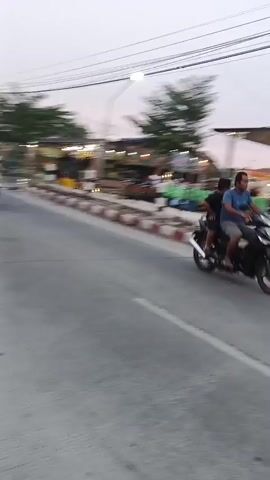 The video captures a street scene with a road in the foreground and a busy outdoor market or food court on the other side of a sidewalk. A few vehicles, including motorcycles, are visible in the market area. People are seated at tables and standing around, suggesting a social gathering or dining experience. There are food stalls and a yellow tent with a sign. The lighting indicates it is either early morning or late evening. The weather appears calm, though a slight breeze might be present due to the movement of the tree.