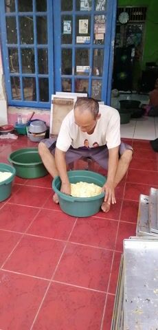 A man is squatting on a red tile floor, in front of a blue door. He is wearing a light-colored t-shirt and dark pants. His hands are in a large green tub filled with a light-colored, lumpy substance, possibly dough or a similar food item. There are two other identical green tubs nearby, one of which also contains the same substance. The man appears to be mixing or preparing the substance. In the background, through the blue door, a green plant can be seen. To the right of the man, there are stacks of metal trays.