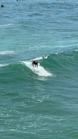 A person in a black wetsuit surfs on a white surfboard in the ocean. The surfer is positioned on the right side of a breaking wave, with their body turned towards the camera. The wave is a vibrant green with white foam cresting. In the background, other waves are visible, as well as a few distant figures in the water. The overall scene depicts a surfing activity in a choppy, blue-green ocean.