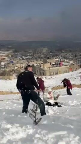 A group of people are playing in the snow on a hill overlooking a town. One man, wearing a black sweater and jeans, is throwing snowballs. Another person is sitting in the snow wearing a white hooded coat. Two other people are also present, one in a red coat and one in dark clothing, both farther up the snowy hill. The town below is a mix of light-colored buildings. The sky is overcast.