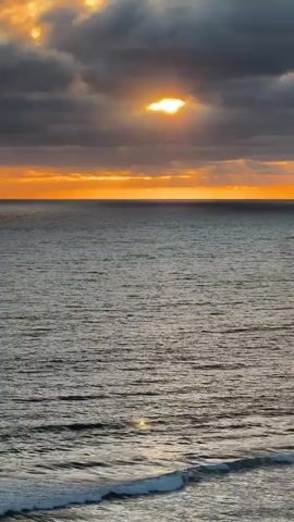 A wide shot captures a dark sky filled with clouds as the sun peeks through a small opening, casting an orange glow. Below the sky, a vast ocean stretches out, its surface a deep gray with gentle waves rolling towards the foreground. The water appears calm with faint ripples and the occasional white crest of a wave. The scene is illuminated by the dim light of the setting or rising sun, creating a dramatic and serene atmosphere. There are no people or objects visible in the frame, only the natural elements of the sky and the sea.