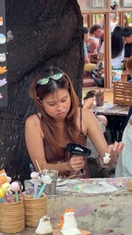 A young woman with reddish-brown hair, wearing a brown tank top and sunglasses perched on her head, is sitting at a table outdoors, appearing to be engaged in a craft activity. She is holding a small white object in her left hand and a tool, possibly a paintbrush or a sculpting tool, in her right hand. She is focused on the object, her facial expression suggesting concentration. In front of her is a table covered in colorful paints and art supplies. To her left, in the foreground, are two woven baskets filled with colorful, pom-pom-like objects on sticks, and two small ceramic cat figurines. Behind her, a large tree trunk dominates the left side of the frame. In the background, several people are visible, some seated at tables, suggesting a cafe or outdoor market setting. The overall lighting indicates it is daytime.