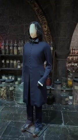 The video displays a dimly lit, atmospheric room that appears to be a potions classroom or laboratory. In the foreground, a mannequin dressed as Professor Severus Snape stands in a dark robe and pants. Behind the mannequin, shelves are filled with numerous glass jars and bottles containing various ingredients, possibly for potions. To the left, a wooden table is set up with more bottles, beakers, and cauldrons, suggesting active experimentation. The walls are made of dark stone, and an arched doorway is visible in the background, further enhancing the magical ambiance. The overall impression is one of a meticulously recreated set from the Harry Potter universe.