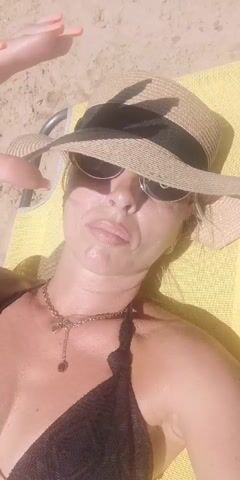 A woman lies on a yellow towel on a beach, wearing a black bikini, sunglasses, and a straw hat. She is holding the brim of her hat with her right hand. Her left arm is partially visible, wearing a blue bracelet. The background consists of sand.