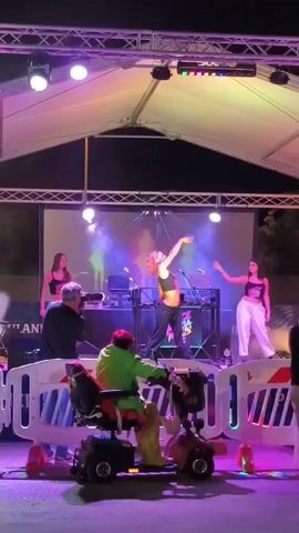 A mobile stage with purple and pink lighting is set up outdoors at night, likely for a performance or event. On the stage, a dancer with red hair in a black crop top and dark pants is performing energetically, bending backward. Two other young women in similar outfits stand on either side of the stage. A DJ booth with a laptop and speakers is visible behind the central dancer. In the foreground, a person with bright red hair, wearing a green jacket and sitting in a motorized scooter, is watching the performance. A photographer in a black jacket is also positioned to the left, capturing the scene. A crowd is gathered behind a temporary white barrier. The atmosphere appears festive with smoke effects adding to the stage lighting.