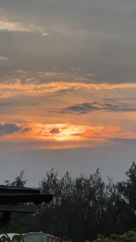 The video shows a sunset with clouds. The sun is partially obscured by clouds, creating a bright orange glow. The clouds are a mix of gray and orange, with some dark patches. Below the clouds, there are trees, and a part of a building can be seen in the lower left corner. The scene is viewed from a slightly elevated position, looking upwards at the sky.