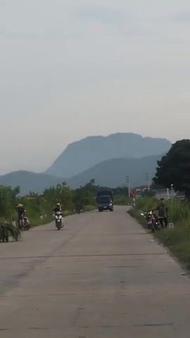 The video shows a two-lane paved road with a truck in the distance. On the left side of the road, there are two people on motorcycles, and on the right side, there is one person standing near a parked motorcycle. In the background, there are hills and trees under a cloudy sky. The lighting suggests it is either early morning or late afternoon.