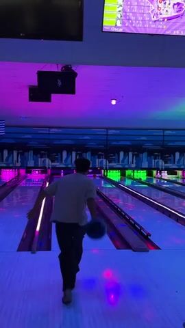 A man is bowling in a dimly lit bowling alley with neon blue and purple lights. He is wearing a light-colored t-shirt and dark pants. He has just released a bowling ball and is watching it roll down the lane. The bowling ball is in motion and heading towards the pins. The bowling alley has multiple lanes with glowing lights and a scoring screen visible in the background. The atmosphere is dark and illuminated by colorful lights.