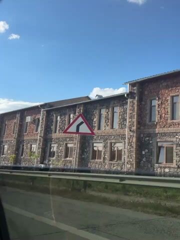 The video captures a drive along a road, likely in Ashtarak, Armenia, given the visible signage. The primary subject is the passing scenery, dominated by a stone building with multiple windows and brick accents. The building appears to be several stories high and stretches across the frame. In the foreground, a metal guardrail separates the road from the area where the building is situated.

The setting is outdoors, under a bright blue sky with scattered white clouds, suggesting fair weather and daytime. The sun is clearly out, casting light and shadows. There are a few small trees and bushes visible near the building, adding a touch of greenery to the otherwise stone and brick landscape. A tall lamppost stands prominently on the left side of the frame in the initial shots.

As the drive continues, the view shifts. We see remnants of stone structures and landscaping on a hill. A blue directional signpost appears, with text in Armenian and English. The Armenian text reads "ԱՇՏԱՐԱԿԻ ՁՈՐ" and below it, "ASHTARAKIDZOR" with an arrow pointing to the right. This confirms the location as Ashtarak Dzōr. Further along, we see power lines and towers, indicative of a more rural or semi-rural environment.

The overall activity is a journey, with the camera positioned inside a vehicle, looking out at the passing landscape. There are no people visible in the footage. The mood is neutral, focusing on the visual experience of the drive and the architectural and natural elements of the location. The time of day appears to be late morning or early afternoon, judging by the sunlight and cloud formations.