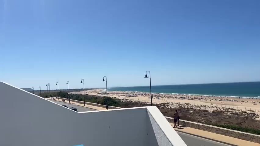 The video pans across a crowded beach on a clear, sunny day. The ocean is a deep blue, with gentle waves reaching the sandy shore. Numerous beachgoers are scattered across the sand, some lying on towels, others walking or playing. Several beach umbrellas provide shade. In the foreground, a white wall frames the view, and a paved walkway runs parallel to the beach. Streetlights with curved tops are spaced along the walkway. A road with a few vehicles is visible behind the walkway. The overall scene depicts a lively and popular beach destination.