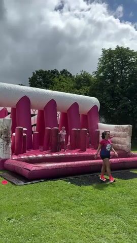 A pink inflatable obstacle course with white beams is set up on a green lawn. Several people, mostly women wearing pink t-shirts, are navigating the course. One person is running across the inflatable surface, kicking up water. Another person, wearing a pink shirt and dark shorts, is standing on the side of the inflatable structure. The sky is cloudy.