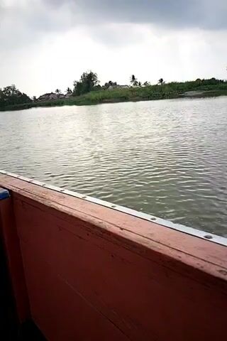 The video begins with a close-up shot of the side of a boat with orange-red paint and metal rivets. The boat is on a body of water, with a cloudy sky overhead. The camera then pans to the left, revealing people sitting inside the boat. The first person visible is wearing a green shirt and a blue headscarf. Next to them, facing forward, is a person in a dark jacket. Further to the right, another person is visible in a light-colored headscarf. The boat has a green canopy overhead, supported by metal poles. The background shows a riverbank with trees and some buildings. The boat appears to be moving.
