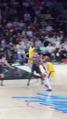 The video shows a basketball game from the perspective of a spectator near the court. A player in a yellow Lakers jersey dribbles the ball as two players in black Miami Heat jerseys guard him. Another player wearing a Lakers jersey approaches. An official in black and white stripes watches. The basketball court is surrounded by spectators. The court has a light-colored wood floor with a graphic design in shades of blue and pink.