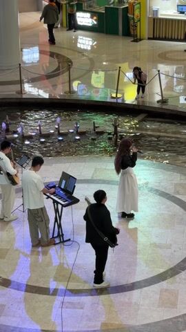 Four people are performing in the center of a shopping mall, near a fountain. One person, dressed in a long white skirt and dark top, is singing into a microphone. To her left, a man plays an acoustic guitar and another man is playing a keyboard. A fourth man, wearing a black suit, stands on the far right of the group, with a guitar. The fountain has small streams of water lit with colorful lights. There are shoppers and other people walking around the mall in the background.  A young girl is standing near the edge of the fountain. The floor is marble or tiled with a circular pattern around the performance area.