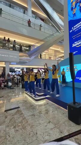 A group of five young performers in yellow shirts and blue pants are dancing on a blue stage with illuminated accents. They are positioned on tiered platforms. Behind them, a crowd of spectators, including adults and children, are watching from a distance. Some spectators are seated on the floor, while others are standing behind a railing. Escalators and multiple levels of the mall are visible in the background, with people observing from the upper floors. A large screen displaying text is partially visible on the right side of the frame. The performance appears to be taking place in a large, well-lit indoor shopping mall.