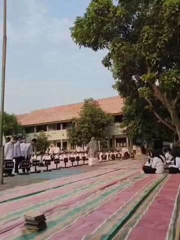 The video shows an outdoor scene at a school, likely during an assembly or event. In the foreground, colorful striped mats are laid out on the ground. A stack of books rests on one of the mats. In the middle ground, a group of students in white uniforms are standing in neat rows, facing a building with an orange roof. Some students are holding black signs with numbers on them, possibly indicating class or group assignments. A person in a grey hijab and white clothing appears to be addressing the students. To the right, another group of students in white uniforms are seated on the ground, facing away from the camera. In the background, the school building is visible, with trees and a clear sky overhead. A flagpole stands to the left of the frame. The overall atmosphere suggests an organized school gathering.