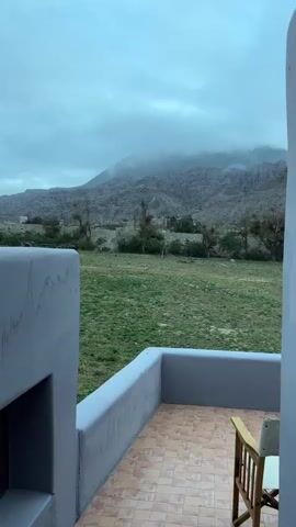 This media file captures a serene view from a patio overlooking a vast, open landscape. In the foreground, a portion of a gray stucco structure with tiled flooring and a simple wooden chair suggests a private dwelling or accommodation. Beyond this immediate setting, a wide expanse of green grass stretches towards a backdrop of rugged, mountainous terrain. The mountains are characterized by their layered, eroded slopes, with some areas obscured by atmospheric haze or low-lying clouds. Several trees, appearing somewhat bare or dry, are scattered across the middle ground, hinting at a possibly arid or cooler climate.

The overall atmosphere is tranquil and somewhat melancholic, due in part to the overcast, cloudy sky and the muted colors of the landscape. The time of day appears to be either early morning or late afternoon, given the soft, diffused light. The weather seems to be cool or overcast, contributing to the subdued mood.

The location is identified as Municipio de Purmamarca, Argentina, a region known for its dramatic geological formations and high-altitude desert landscapes. The scene evokes a sense of remoteness and natural beauty, inviting contemplation. No people are visible in the media, and no text is present. The focus is entirely on the natural environment and the tranquil atmosphere it provides.