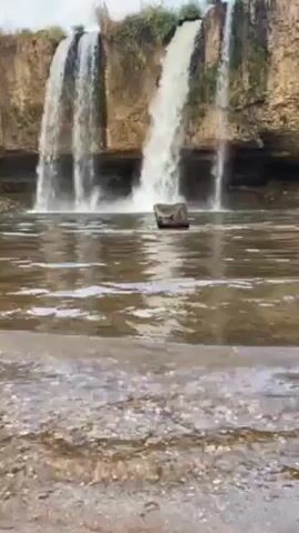The video shows a picturesque waterfall cascading over a rocky cliff into a shallow pool. Two distinct streams of water plunge downward, creating a refreshing and scenic view. The water is somewhat murky, reflecting the sky and surrounding environment. A dark, submerged object is visible in the pool, adding a touch of mystery to the scene. The sky is partly cloudy, contributing to the overall serene and natural setting.