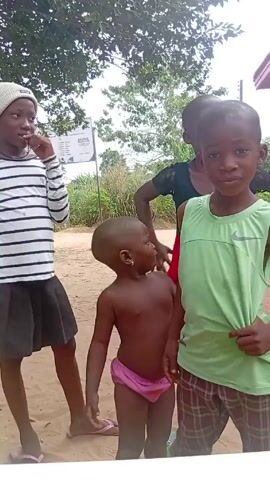 A video captures four children in an outdoor setting. The first child, a girl, stands to the left, wearing a black and white striped long-sleeved shirt, a black skirt, a white beanie, and pink flip-flops. She has her hand to her face. A smaller child stands in the center, only wearing pink underwear and pink flip-flops. To the right is another child, a boy with a light green Nike tank top and brown plaid shorts, smiling. Behind them is an adult woman. They are standing on a dirt ground with greenery and trees in the background. A sign is slightly visible. The children appear to be posing for the camera.