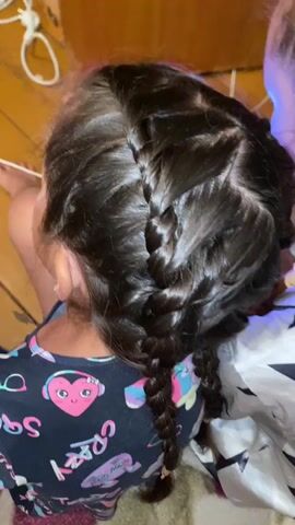 The video shows a close-up view of the back of two young girls' heads. The girl in the foreground has dark brown hair styled in braids, with the back of her head and braids filling most of the frame. She is wearing a dark shirt with a colorful graphic design on it. To the right, another girl is visible, also with dark brown hair, wearing a white pajama top with black stars. She is holding a smartphone, illuminated by its screen, and appears to be looking at it. The background is dimly lit and shows what appears to be a wooden floor and possibly a rug. The overall impression is that the girls are sitting or lying down together, possibly indoors.
