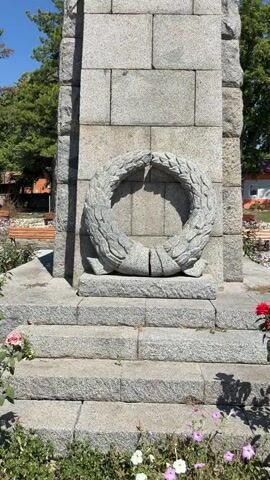 The video shows a close-up, upward-angled view of a stone monument. The monument has a wreath carved into its lower section, and above that, a star is visible. At the very top, a statue of a person holding a rifle and raising their other hand stands against a clear blue sky. Trees and some buildings are visible in the background.