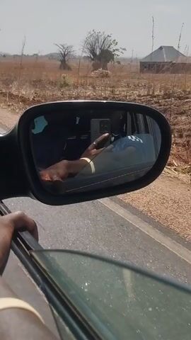 The video shows a person in a car, possibly driving on a rural road. The view is from inside the car, with the driver visible in the side mirror. The landscape outside is dry and grassy with scattered trees and a clear sky. The person holding the camera is partially visible, with their arm and hand near the car window. There's a noticeable wind sound, likely due to the vehicle's speed.