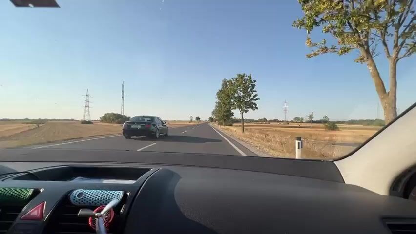 The video begins with a view from inside a car, looking out through the windshield. The dashboard is partially visible in the foreground, with a small white sticker on the top left. The car is driving on a paved road with a clear blue sky overhead. Fields of dry grass and crops are visible on either side of the road. In the distance, a black car is driving ahead on the same road. There are power lines and utility poles visible in the background. As the video progresses, the car continues to drive, and the perspective shifts slightly, showing more of the passenger side of the car and the rearview mirror. The fields and trees continue to pass by. The overall impression is of a car journey through a rural landscape on a sunny day.