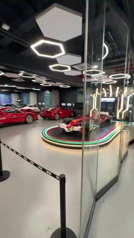 The video shows an indoor display of several red sports cars. In the foreground, a Formula 1 race car is prominently displayed on a circular platform with green and red lighting. Further back, a line of red sports cars is parked, some with their doors open. The environment appears to be a showroom or a museum, with a dark ceiling featuring hexagonal lighting fixtures and a clean, grey floor. The cars are separated by stanchions and black and white striped ropes.